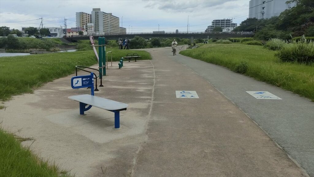川沿いのジョギングコースと、複数の健康遊具が設置された広場の風景。市街地近くの緑道で人々が運動している
