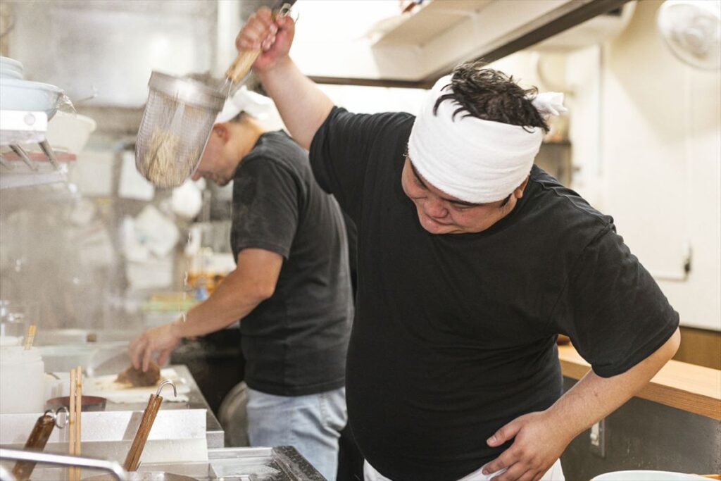 ラーメン店の厨房で湯切り作業を行う職人。蒸気が立ち込める中、汗をかきながら働く姿が熱中症リスクを物語る。