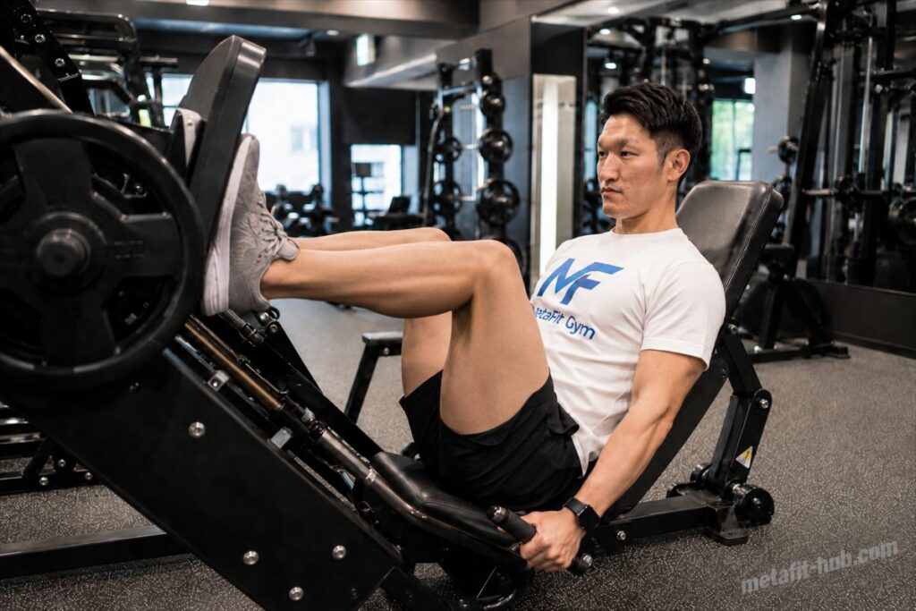 ジムのレッグプレスマシンで大腿四頭筋を鍛えるトレーニーの男性。MetaFit GymのTシャツを着て集中した表情でプレスしている。
